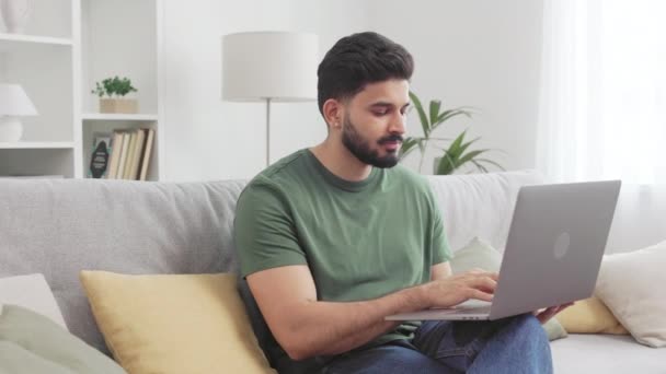 Ελκυστικός νεαρός που κάθεται σε αναπαυτικό καναπέ χρησιμοποιώντας οικιακό internet ενώ εργάζεται σε ασύρματο φορητό υπολογιστή στο σπίτι. Ινδουιστής ελεύθερος επαγγελματίας σε casual ενδυμασία με χαρούμενη διάθεση απολαμβάνοντας απόσταση εργασίας. - Πλάνα, βίντεο