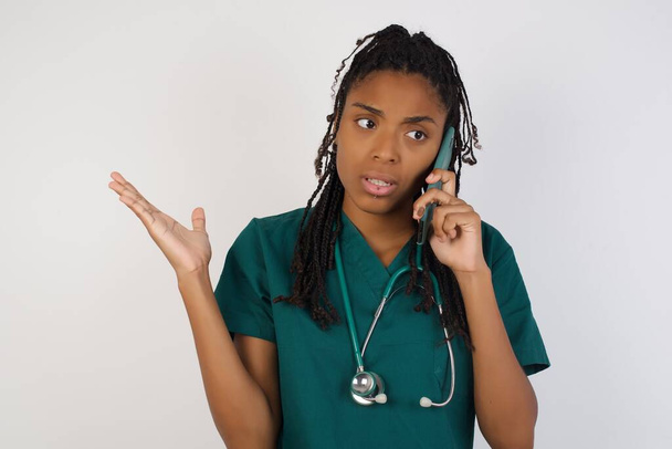 Молода афроамериканська жінка-лікар розмовляє по телефону над ізольованим тлом, напруженим рукою на обличчі, шокована соромом і несподіваним обличчям, розлючена і розчарована. Страх і засмучення за помилку. - Фото, зображення