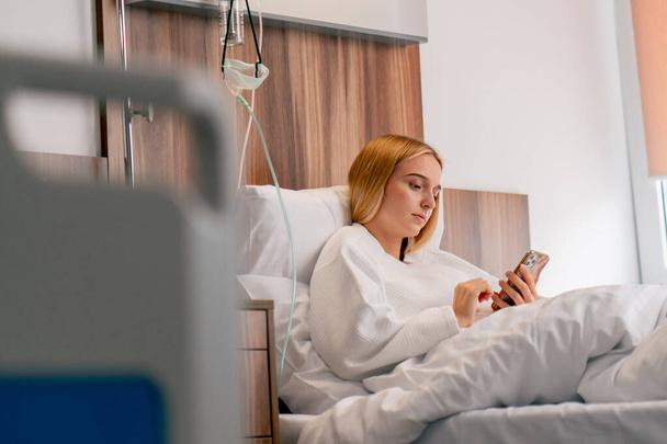 Uma menina triste em um roupão de banho encontra-se em um quarto de hospital e, infelizmente, olha para o telefone enquanto espera por seu médico - Foto, Imagem
