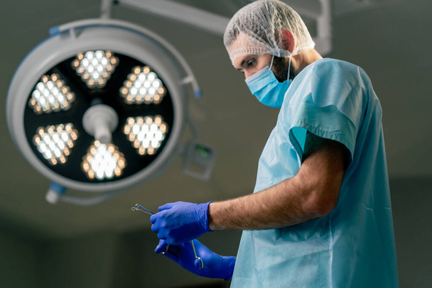 Un médico en el quirófano con un uniforme protector y un tocado especial se pone guantes antes de una operación en un quirófano - Foto, imagen
