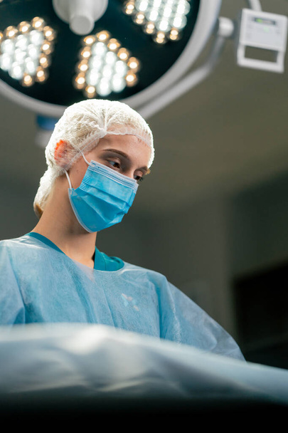 Девушка-ассистент хирурга в форме и медицинской маске передает скальпель своему коллеге во время операции - Фото, изображение