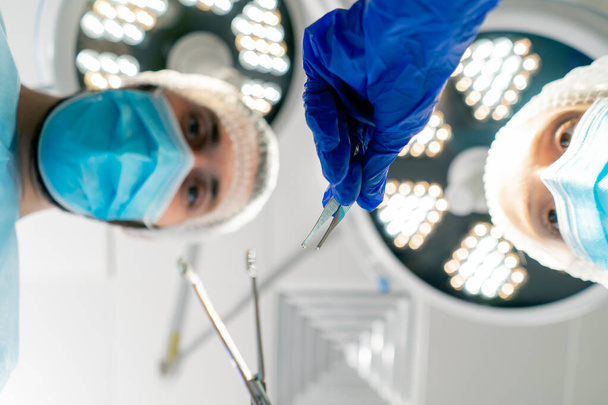 Επαγγελματίες χειρουργοί με στολή στο φόντο των λαμπτήρων λυγισμένα πάνω από έναν ασθενή που βρίσκεται στο χειρουργικό τραπέζι για να εκτελέσει μια λειτουργία - Φωτογραφία, εικόνα