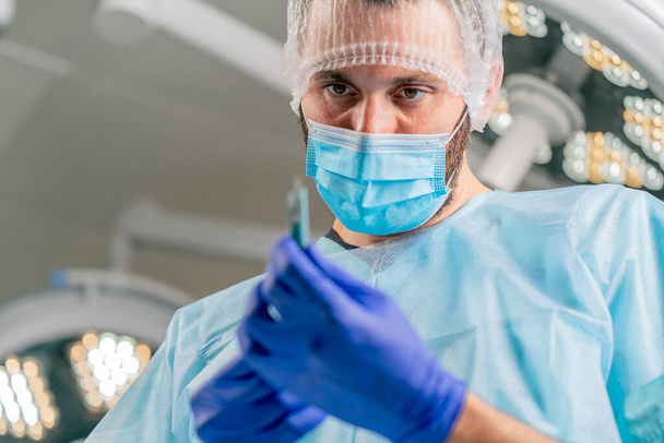 Gros plan d'un chirurgien dans une coiffe protectrice et un masque médical tenant un scalpel dans ses mains et le regardant attentivement - Photo, image