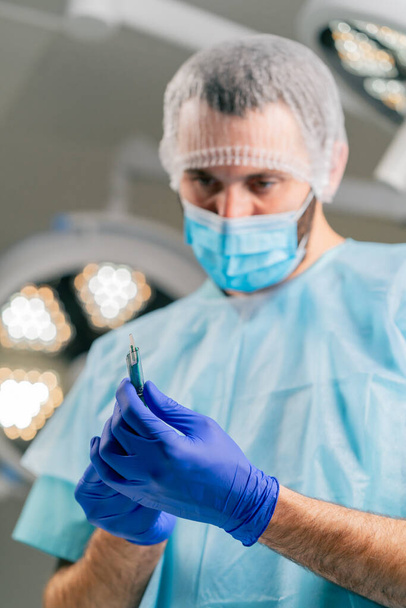 Крупный план хирурга в защитном головном уборе и медицинской маске, держащего скальпель в руках и внимательно рассматривающего его - Фото, изображение