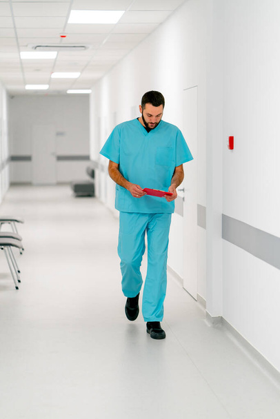 Високий чоловічий лікар з бородою йде по лікарняному коридору з папкою документів - Фото, зображення