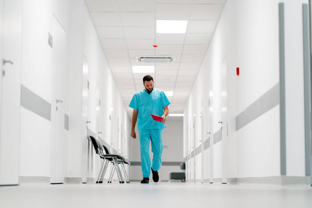 Een lange mannelijke dokter met een baard loopt door de ziekenhuisgang met een map van documenten - Foto, afbeelding