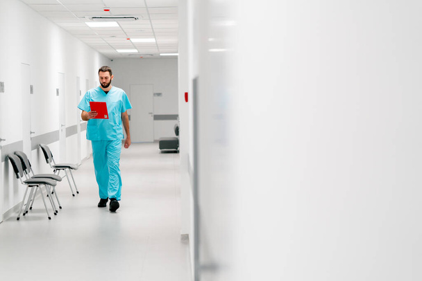 Een lange mannelijke dokter met een baard loopt door de ziekenhuisgang met een map documenten en komt zijn kantoor binnen - Foto, afbeelding