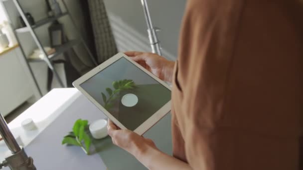 Medium close-up over-shoulder shot of unrecognizable female blogger holding tablet computer in hands, focusing on jar of cream and green leaf on table, and taking photos - Footage, Video