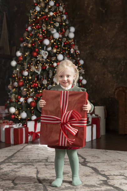Cute little girl with big red gift box. Smiling blonde child near Christmas tree. Christmas Eve. - Foto, afbeelding