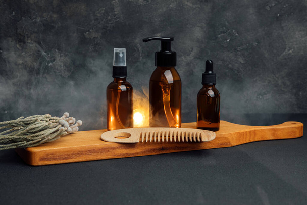 Borostyánsárga palackok arckozmetikumokkal egy fa tálcán, száraz ágakkal sötét alapon, arcszérummal. Előnézet - Fotó, kép