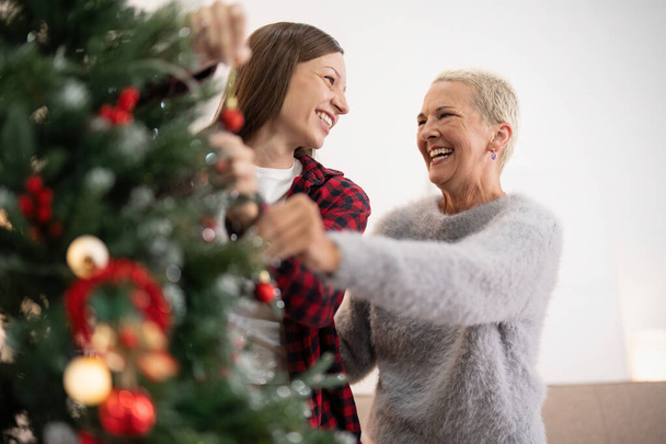 Familie wintervakantie en concept gelukkig senior vrouw met volwassen dochter versieren kerstboom thuis. - Foto, afbeelding