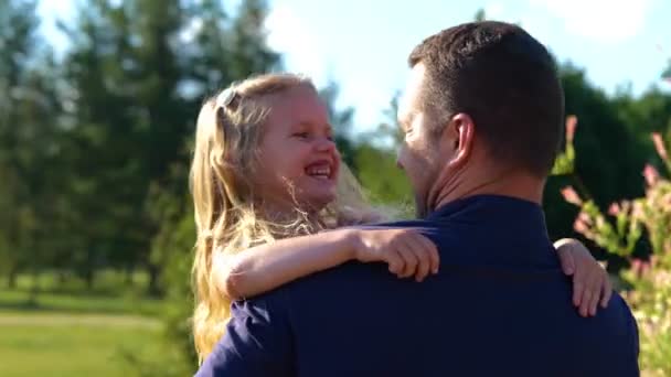 Zadní část mladého otce drží svou dcerku a usmívá se. Detailní záběr hezkého kluka s tátou venku v parku za slunečného dne. Rodinný čas. V reálném čase. Šťastné dětství. Veselá dívka s rodičem - Záběry, video
