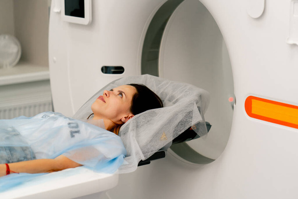 Ένα κορίτσι σε ένα ιατρικό φόρεμα βρίσκεται σε ένα κινητό καναπέ ενός μηχανήματος απεικόνισης μαγνητικού συντονισμού και προετοιμάζεται για μια διάγνωση - Φωτογραφία, εικόνα