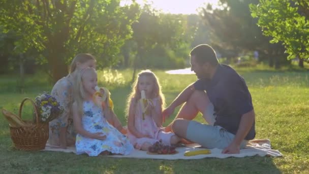 Blanke familie met leuke blonde meisjes in het park in de zomer. Gelukkige kinderen eten fruit zittend op een deken buiten. Echte tijd. Gelukkige jeugd. Familieweekend. Kinderen eten bananen met ouders - Video