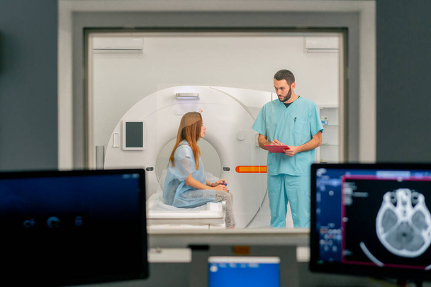 Számítógépes tomográfiás vizsgálat előtt a beteggel kommunikáló orvos hátterében diagnosztikai monitorok - Fotó, kép