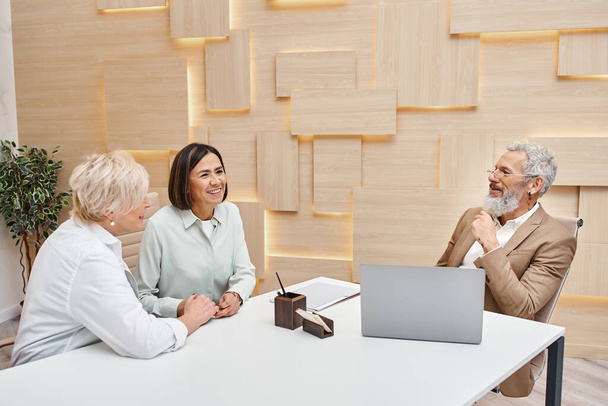 middelbare leeftijd interraciale lgbt paar bespreken onroerend goed in de buurt van bebaarde makelaar in onroerend goed kantoor - Foto, afbeelding