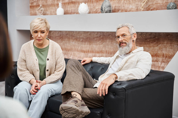 pareja de mediana edad sentada en un sofá de cuero y mirando a un consejero familiar durante la consulta - Foto, imagen