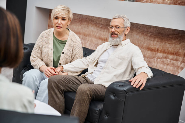 μεσήλικες ζευγάρι κάθεται σε δερμάτινο καναπέ και ψάχνει σε ψυχολόγο κατά τη διάρκεια της οικογενειακής συμβούλων - Φωτογραφία, εικόνα