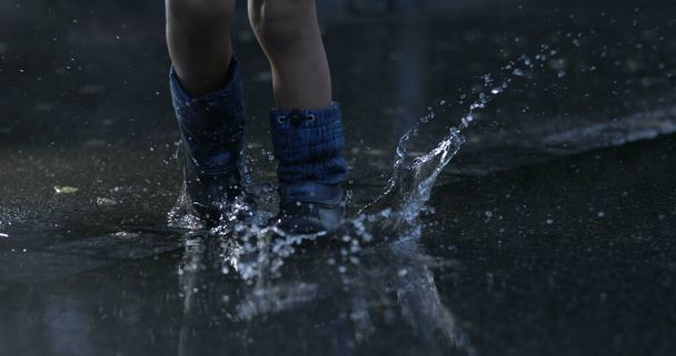 Ребенок шагает в водяной луже в супер замедленной съемке в дождевых сапогах, брызгающих капельками жидкости повсюду, заснятых высокоскоростной камерой на 800 кадров в секунду - Фото, изображение