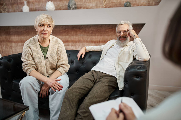 mujer de mediana edad sentada en el sofá de cuero cerca de marido sonriente durante la sesión de terapia familiar - Foto, imagen
