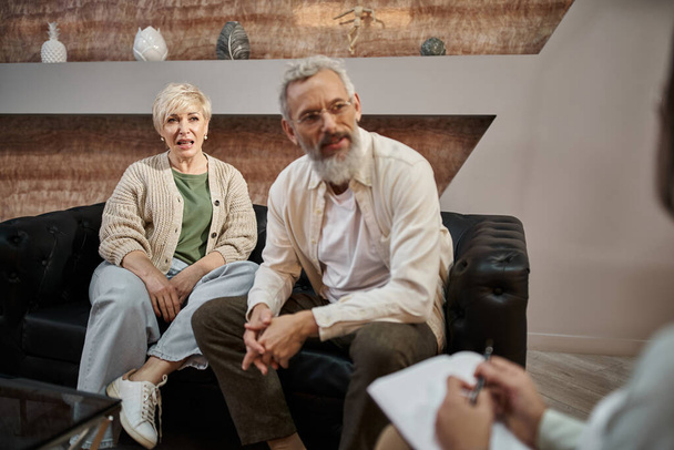 emotional middle aged couple sitting on couch and talking to psychologist during therapy session - Photo, Image