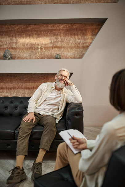 χαρούμενος γενειοφόρος μεσήλικας άνδρας με τατουάζ κάθεται σε δερμάτινο καναπέ και ψάχνει σε ψυχολόγο - Φωτογραφία, εικόνα