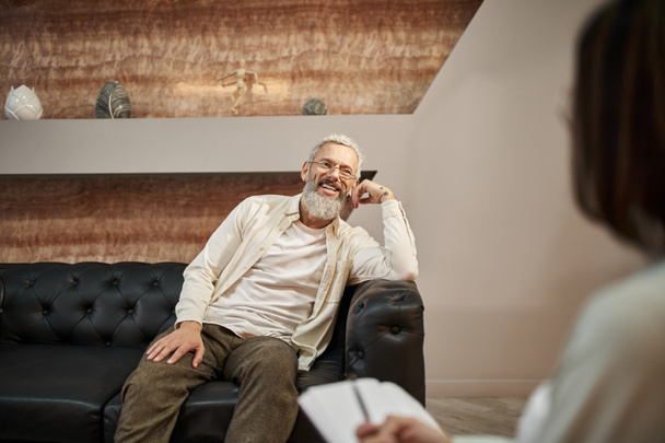 alegre barbudo hombre de mediana edad con tatuaje sentado en el sofá de cuero y mirando psicólogo - Foto, imagen