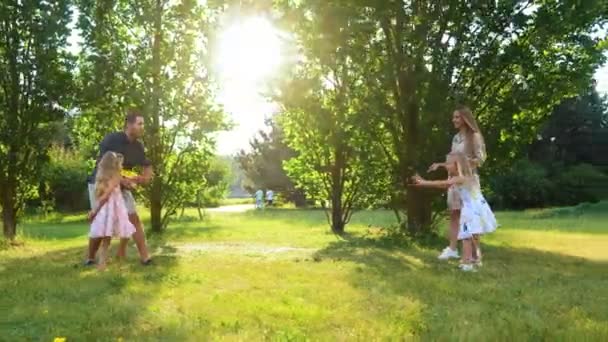 Happy positive family throwing frisbee disk in park. Smiling girl catching frisbee plate. Joyful parents playing frisbee with daughters. Family enjoying summer outdoor activity. Real time. Happy - Footage, Video
