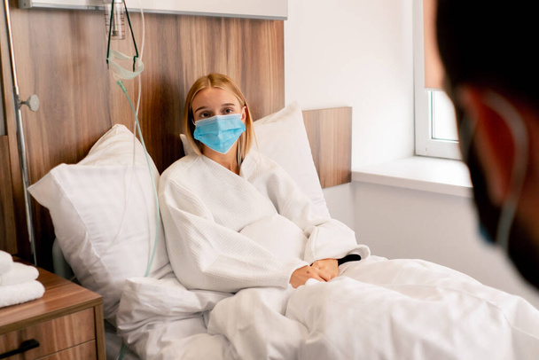 Ένα κορίτσι σε ένα δωμάτιο νοσοκομείου με μια ιατρική μάσκα σε καραντίνα περιγράφει σε ένα γιατρό παράπονα για την υγεία της - Φωτογραφία, εικόνα