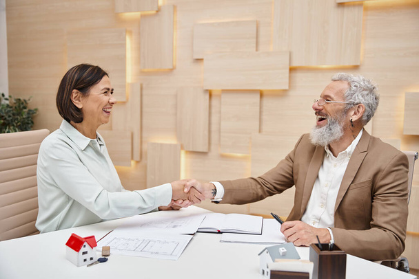 счастливая женщина улыбается и пожимает руку риелтору среднего возраста в офисе недвижимости, продажа недвижимости - Фото, изображение