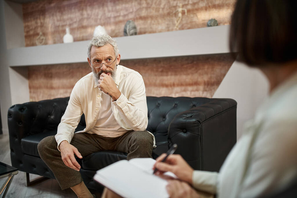 обеспокоенный бородатый мужчина среднего возраста с татуировкой сидя на кожаном диване и глядя на психолога - Фото, изображение