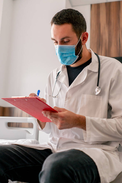 Tıbbi maske ve hastane önlüğü takmış bir doktorun tıbbi geçmişine hastanın şikayetlerini yazdığı yakın plan bir fotoğraf. - Fotoğraf, Görsel