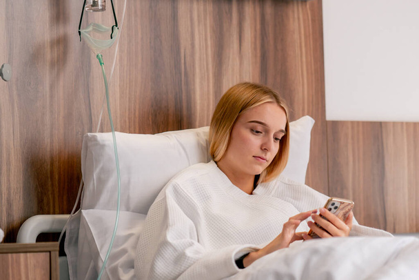 Surullinen tyttö kylpytakissa makaa sairaalahuoneessa ja valitettavasti katsoo puhelintaan odottaessaan lääkäriään. - Valokuva, kuva