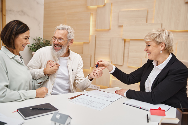 middle aged realtor giving key from new house to happy married couple in real estate office - Photo, Image