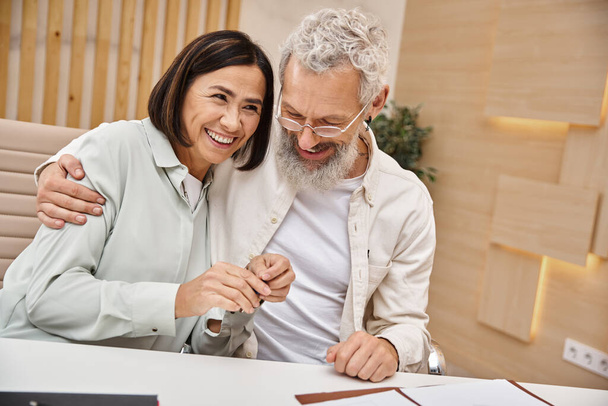 happy bearded man hugging multiracial wife holding key from new house in real estate office - Photo, Image