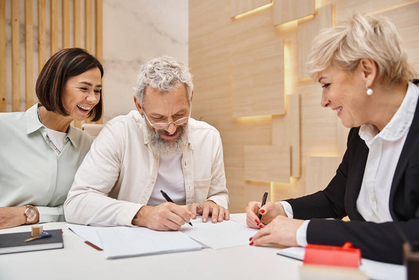 gelukkige man van middelbare leeftijd tekenen contact in de buurt van vrouw en gelukkig makelaar in kantoor, nieuw huis - Foto, afbeelding