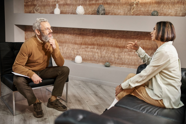 Бородатый психолог средних лет в очках разговаривает с клиенткой во время сеанса терапии - Фото, изображение