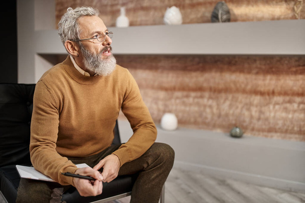 szakállas középkorú pszichológus szemüvegben toll mellett notebook és hallgatni ülés közben - Fotó, kép