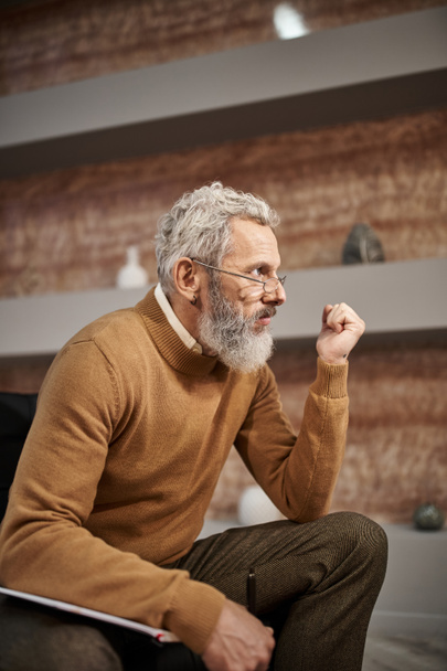 bärtiger Psychologe mittleren Alters mit Brille, Stift neben Notizbuch haltend und während der Sitzung zuhörend - Foto, Bild