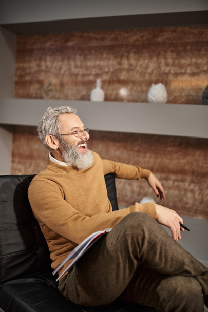 szczęśliwy psycholog w średnim wieku w okularach śmiejący się i siedzący na fotelu podczas sesji terapeutycznej - Zdjęcie, obraz