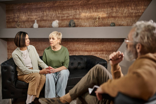 Психолог средних лет в очках слушает разговоры обеспокоенной лесбийской пары во время сеанса - Фото, изображение