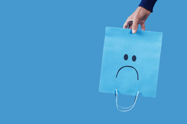 un hombre sostiene una bolsa de papel azul, con una cara triste pintada en ella, vacía boca abajo sobre un fondo azul, con un poco de espacio en blanco a la izquierda - Foto, imagen