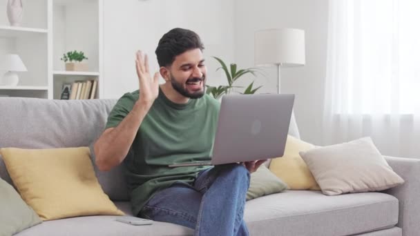 Glimlachende man met baard zwaaiend met de hand voor begroeting gesprekspartner tijdens videoconferentie op moderne laptop. Aantrekkelijke indiaanse man in casual kleding zitten op de bank en genieten van online communicatie. - Video