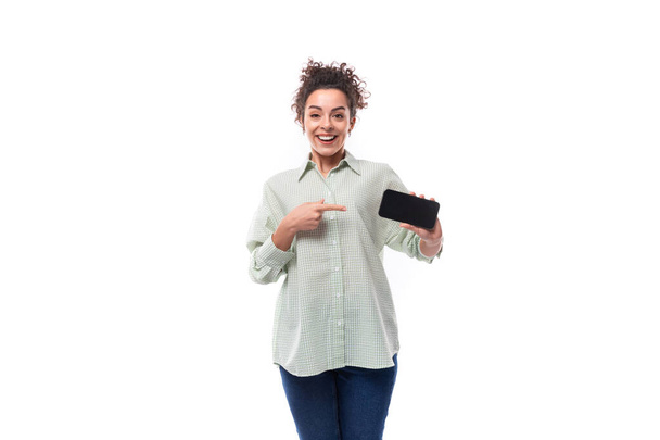 hezká mladá černosrstá kudrnatá žena s neopatrnou stylovou houskou v košili radostně ukazuje obrazovku telefonu vodorovně. - Fotografie, Obrázek