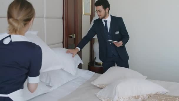 Au-dessus de la vue d'épaule de la femme de ménage mettant des oreillers et faisant le lit dans la chambre d'hôtel avec le directeur en costume - Séquence, vidéo
