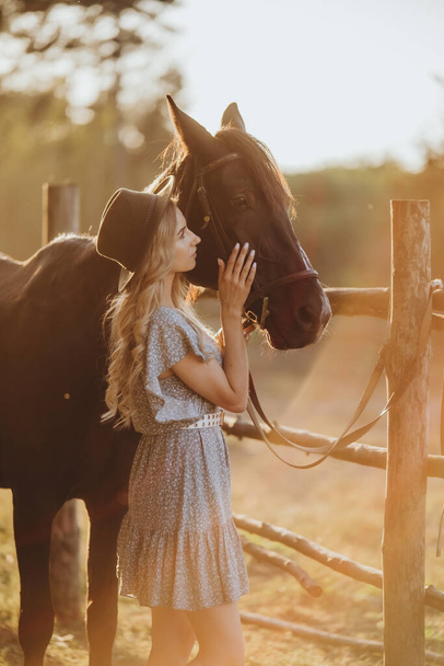 A young woman with a horse stands near the fence on her ranch at sunset in the fall. Breeding thoroughbred horses for equestrian sport. - Photo, Image