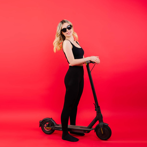 Hermosa mujer joven emocional en una ropa deportiva en scooter eléctrico sobre fondo rojo y blanco - Foto, Imagen
