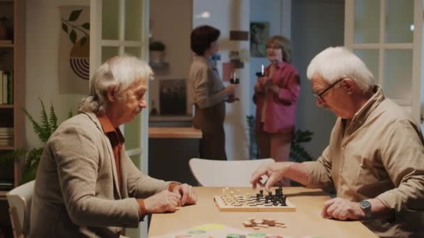 Средний снимок концентрированных пожилых мужчин, играющих в шахматы, в то время как их жены пьют вино на заднем плане на домашней вечеринке - Кадры, видео