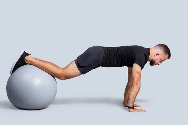 Serious muscular young european guy with beard, make push-ups exercises with fit ball for hands, enjoy workout, isolated on gray studio background. Active training, body care - Photo, Image