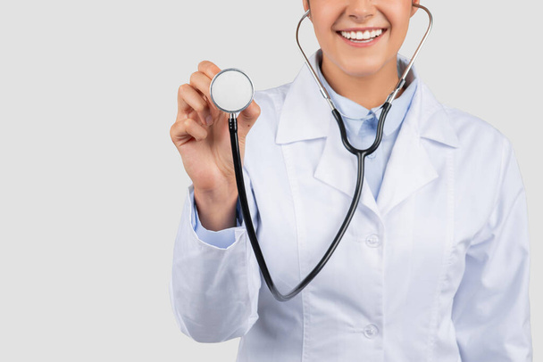 Hymyilevä eurooppalainen tuhatvuotinen lääkäri valkoisessa takissa, kuuntele hengittää stetoskoopilla, rajataan, eristetään harmaalla studiotaustalla, lähikuva. Lääkärintarkastus, terveyspalvelut - Valokuva, kuva